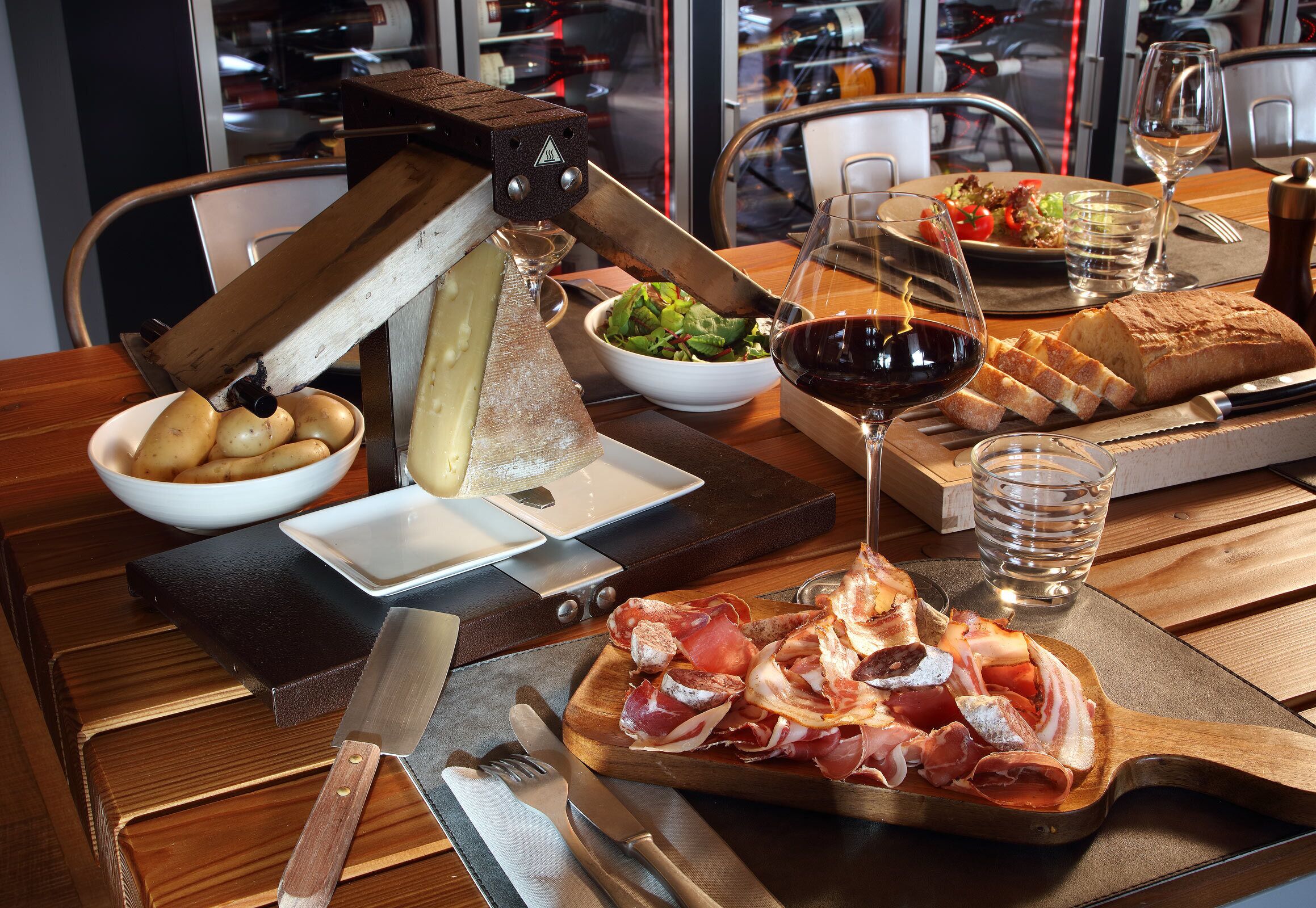 A wooden table with a cheese slicer, a bowl of potatoes, a plate of prosciutto, a loaf of bread, a glass of red wine, and a glass of water.
