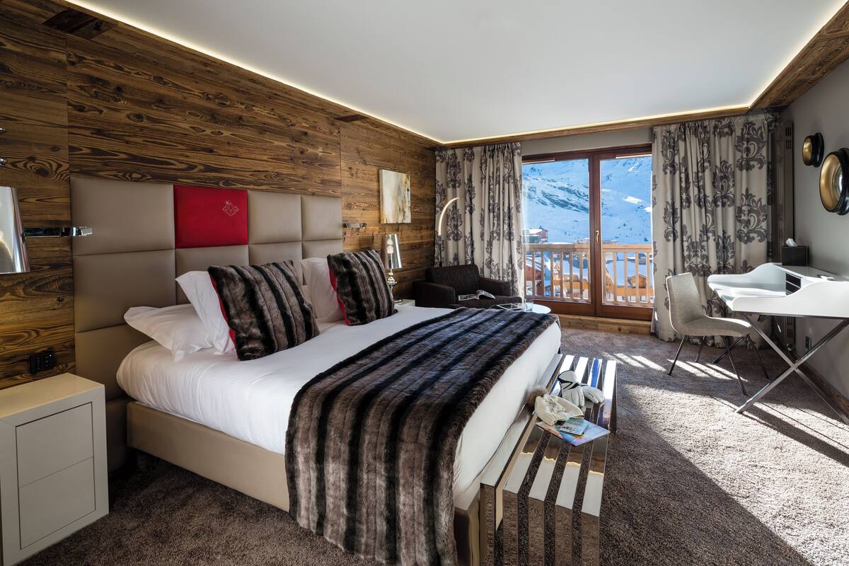 Habitación de hotel moderna con pared de madera, una cama grande con manta de piel, un escritorio con silla y vistas a la montaña a través de la ventana.