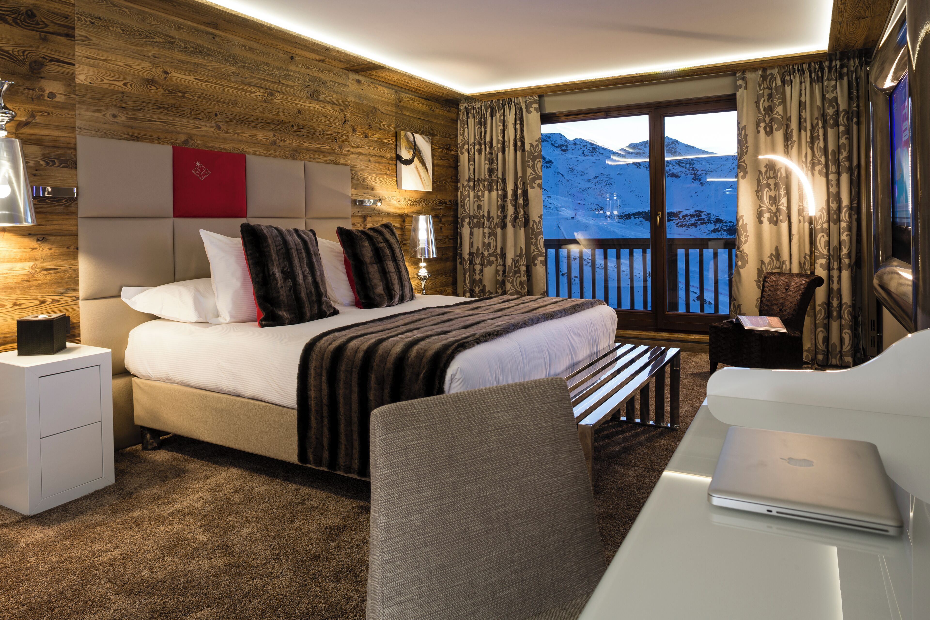 A modern hotel room with a large bed, a desk with a laptop, and a view of a snowy mountain.