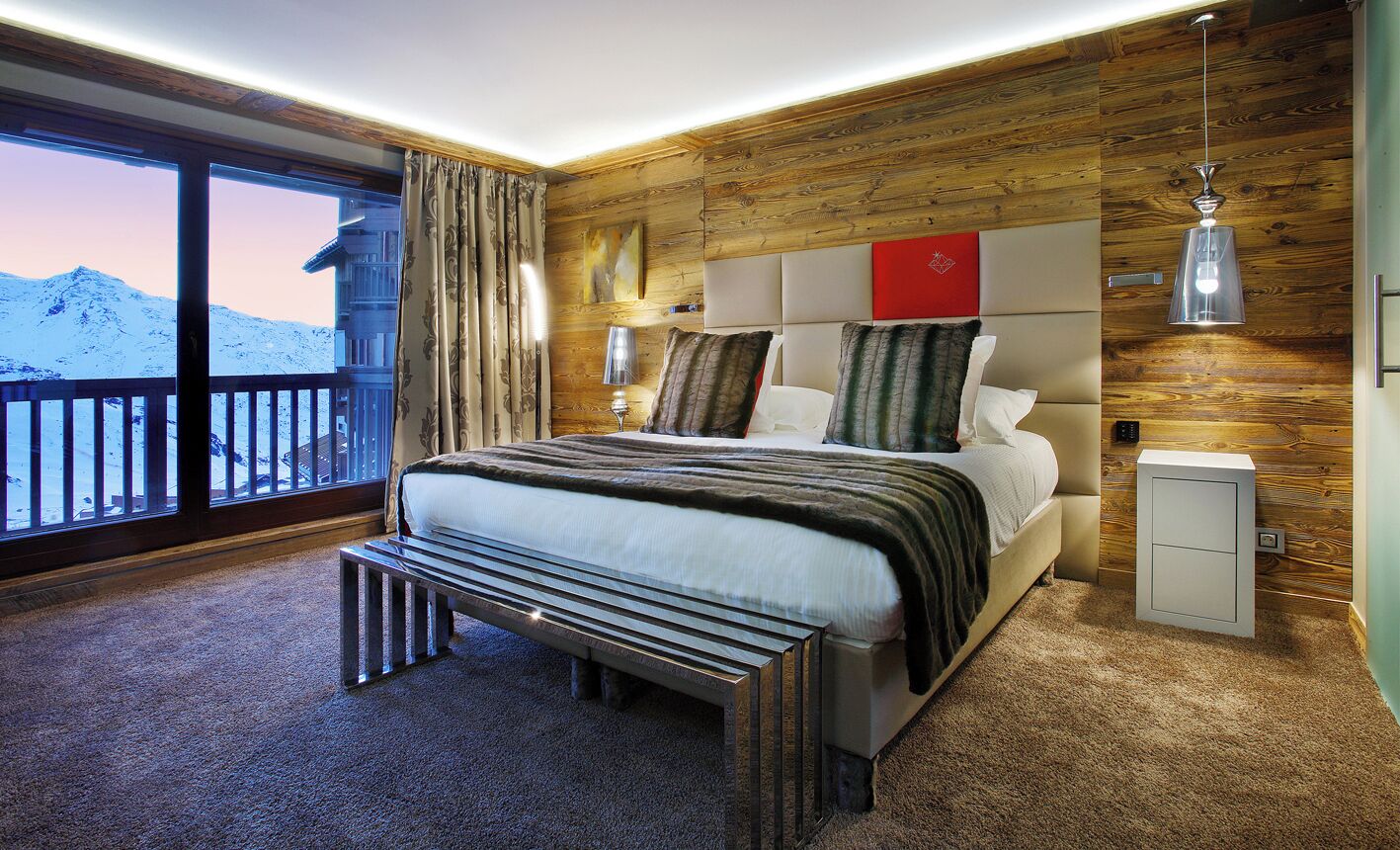 Una habitación de hotel moderna con una cama grande, paneles de madera, una mesita de noche y vistas a montañas nevadas.
