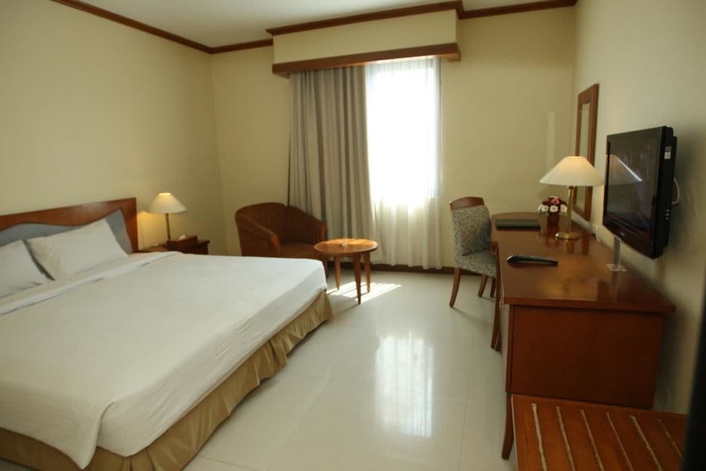 A hotel room with a large bed, a desk with a TV, a chair, a small table, and a window with curtains.