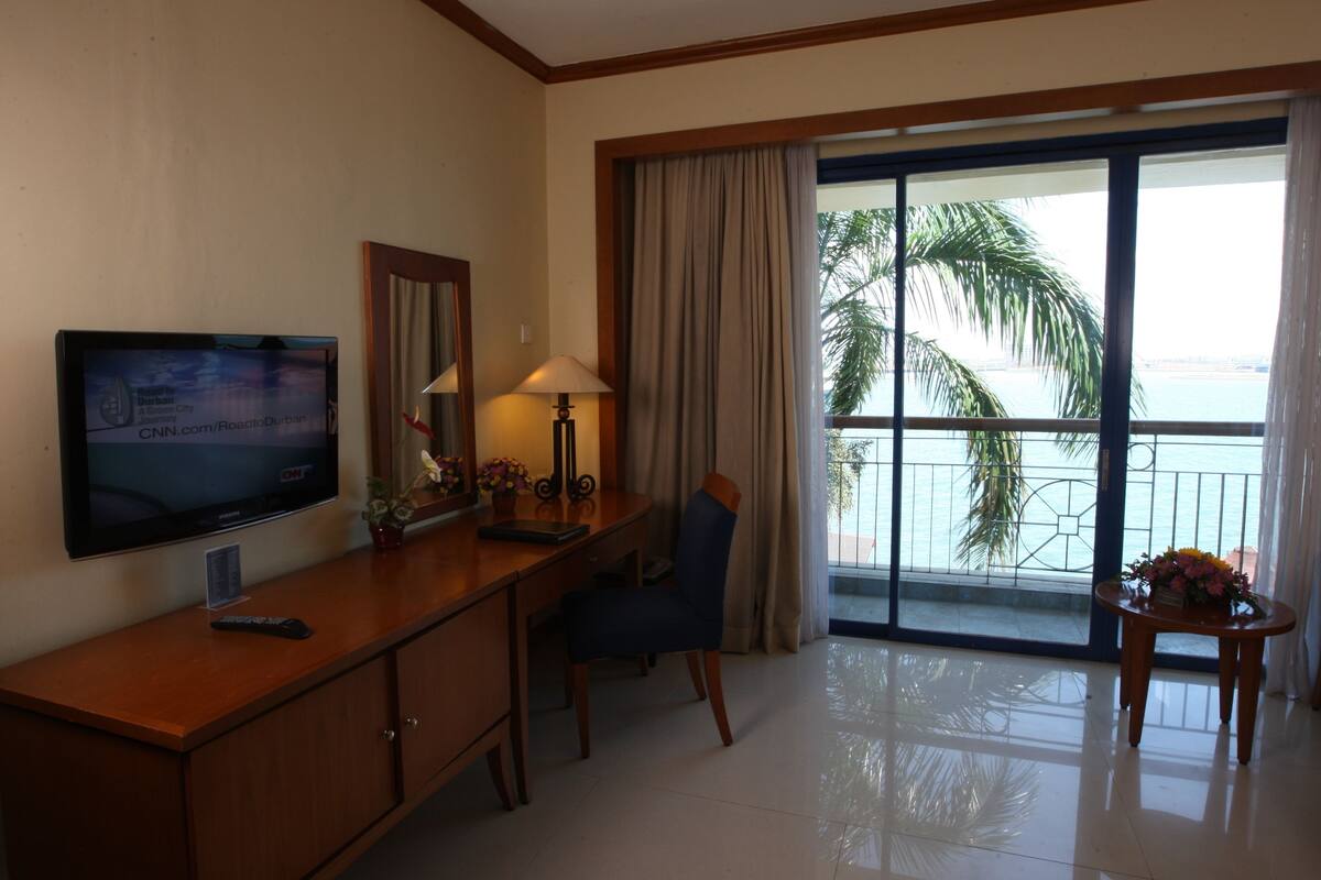 Une chambre d’hôtel avec un téléviseur posé sur une console en bois, un bureau avec une lampe, une chaise et une vue sur des palmiers par une grande fenêtre.