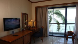 Habitación de hotel con televisor sobre una consola de madera, un escritorio con lámpara, una silla y vista a palmeras a través de un ventanal grande.