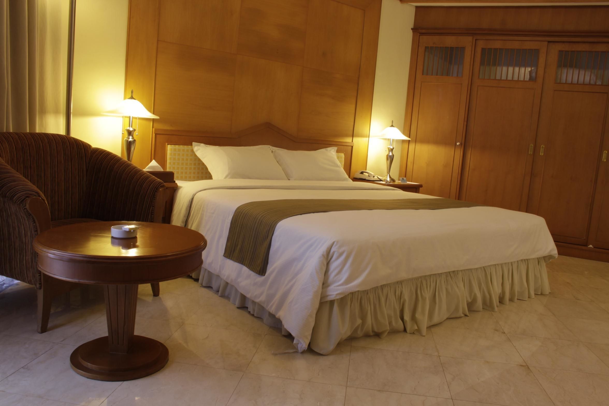 A hotel room with a double bed, a round wooden table, two bedside lamps, a wooden wardrobe, and a striped armchair.