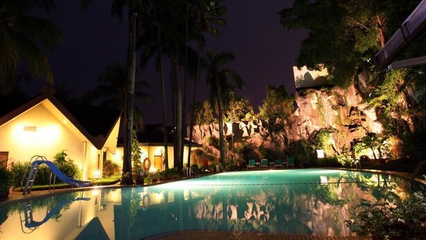 Zona de piscina nocturna en un resort, con piscina cristalina, palmeras, tobogán y edificio con fachada iluminada.