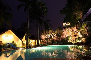 A nighttime resort pool area with a clear pool, palm trees, a slide, and a building with a lighted facade. - Makassar Golden Hotel (Makassar)