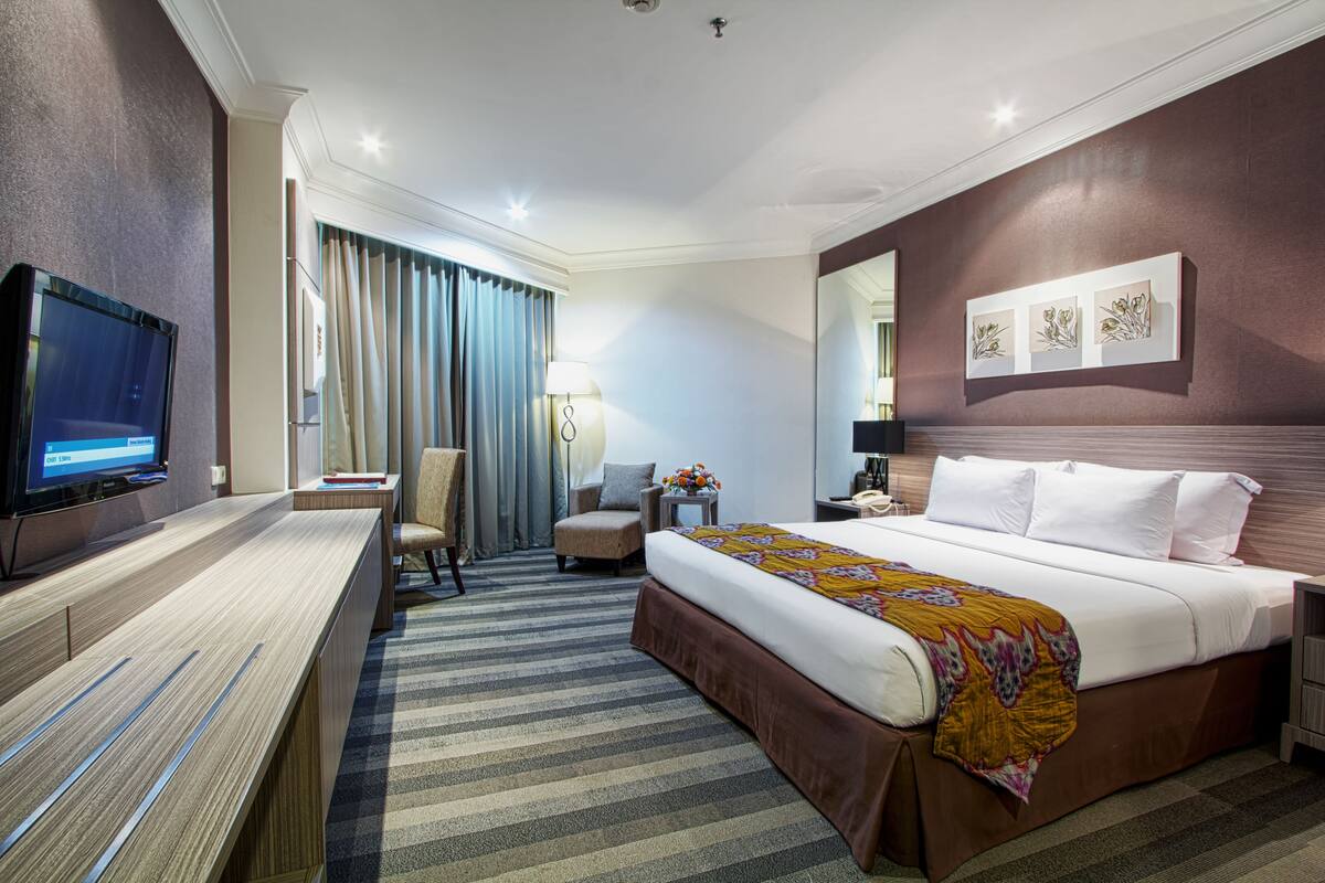 A hotel room with a large bed, a desk with a flat-screen TV, a chair, a nightstand, and a wall-mounted artwork.