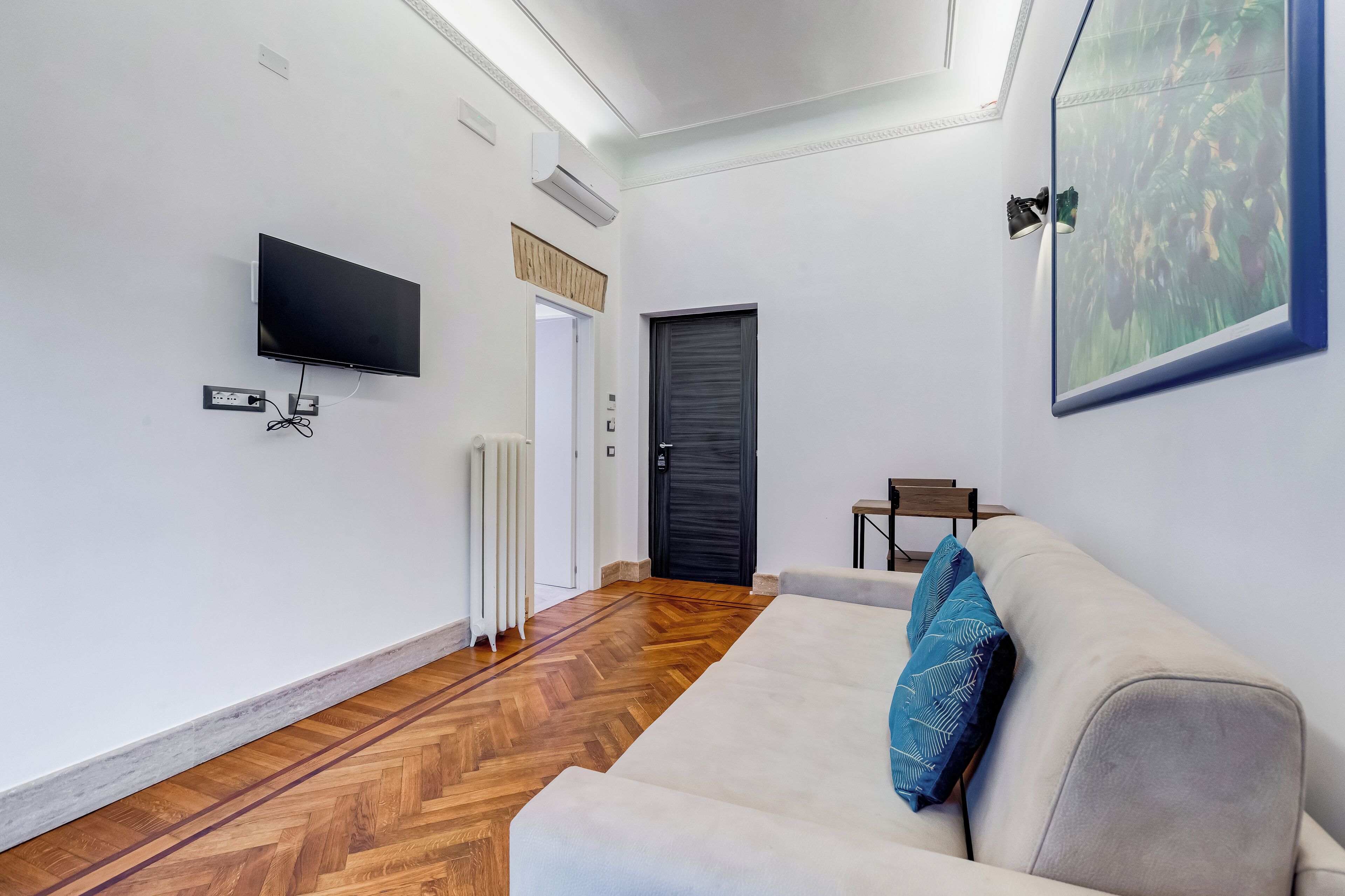 A modern living room with a sofa, a flat-screen TV mounted on the wall, a radiator, and a door.