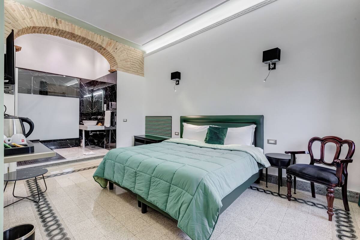 A bedroom with a green bed, a wooden chair, a small table, a mirror, and a wall-mounted light fixture.