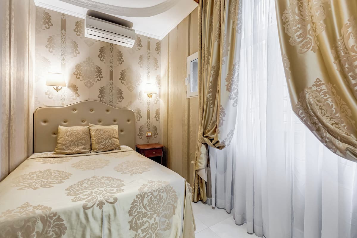 A hotel room with a large bed, two wall-mounted lamps, an air conditioner, and a window with curtains.