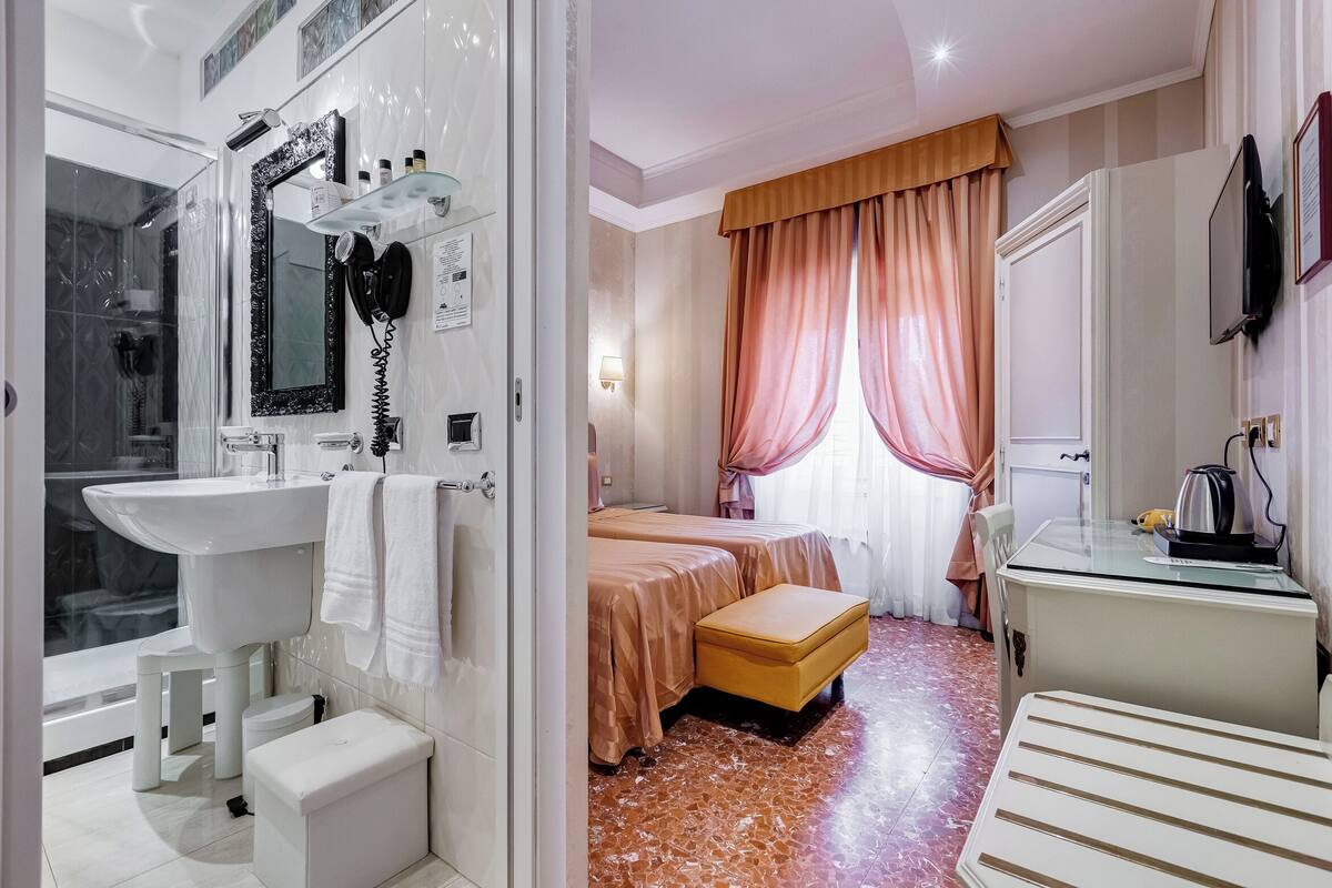 A bathroom with a sink, mirror, and shower. There is a bed with a bench and a TV on a dresser.