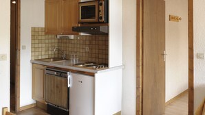 A compact kitchen with wooden cabinets, a microwave, a stove, and a dishwasher. There is a dining table with chairs in the foreground. - La Résidence (La Clusaz)