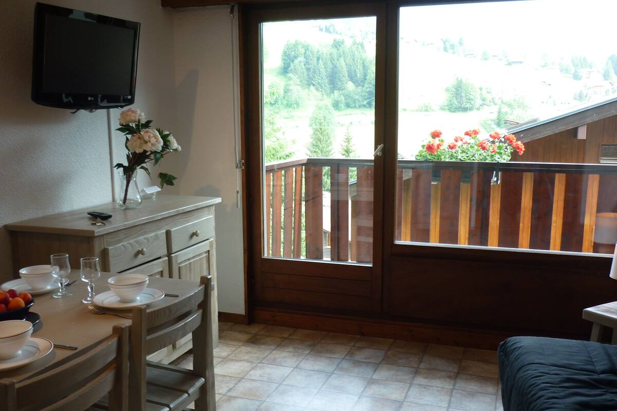 Un coin repas avec une table dressée pour deux personnes, un téléviseur fixé au mur et un balcon doté d’une balustrade en bois et de fleurs.
