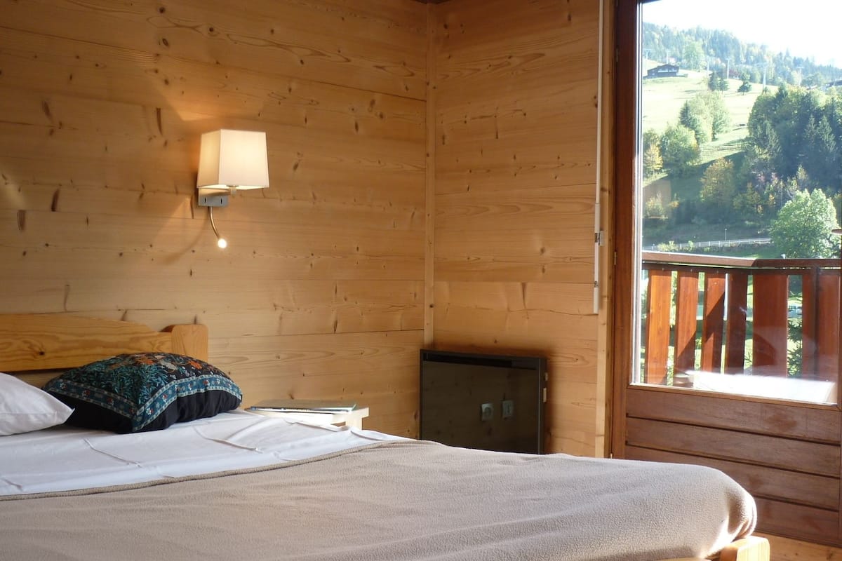 Une chambre rustique en bois, avec un lit, une table de chevet, une lampe et une fenêtre donnant sur un paysage boisé.