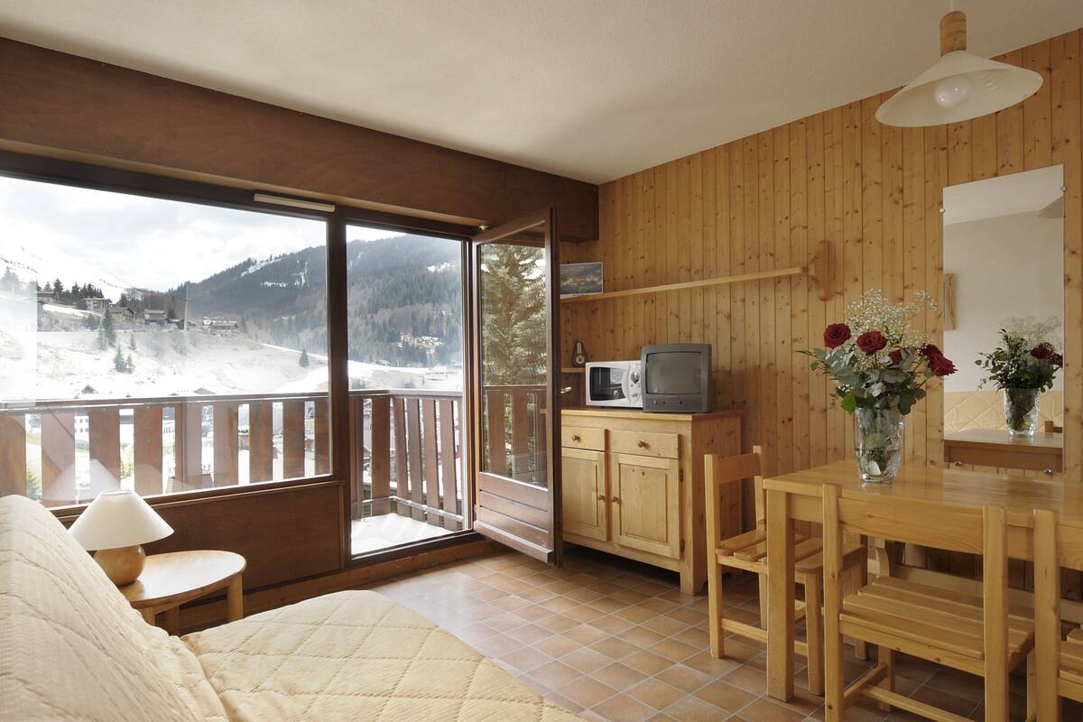 Une pièce chaleureuse avec un décor intérieur en bois, un lit, une petite table avec une lampe, une table à manger avec des chaises, un four à micro-ondes et une vue sur un paysage enneigé par une grande fenêtre.