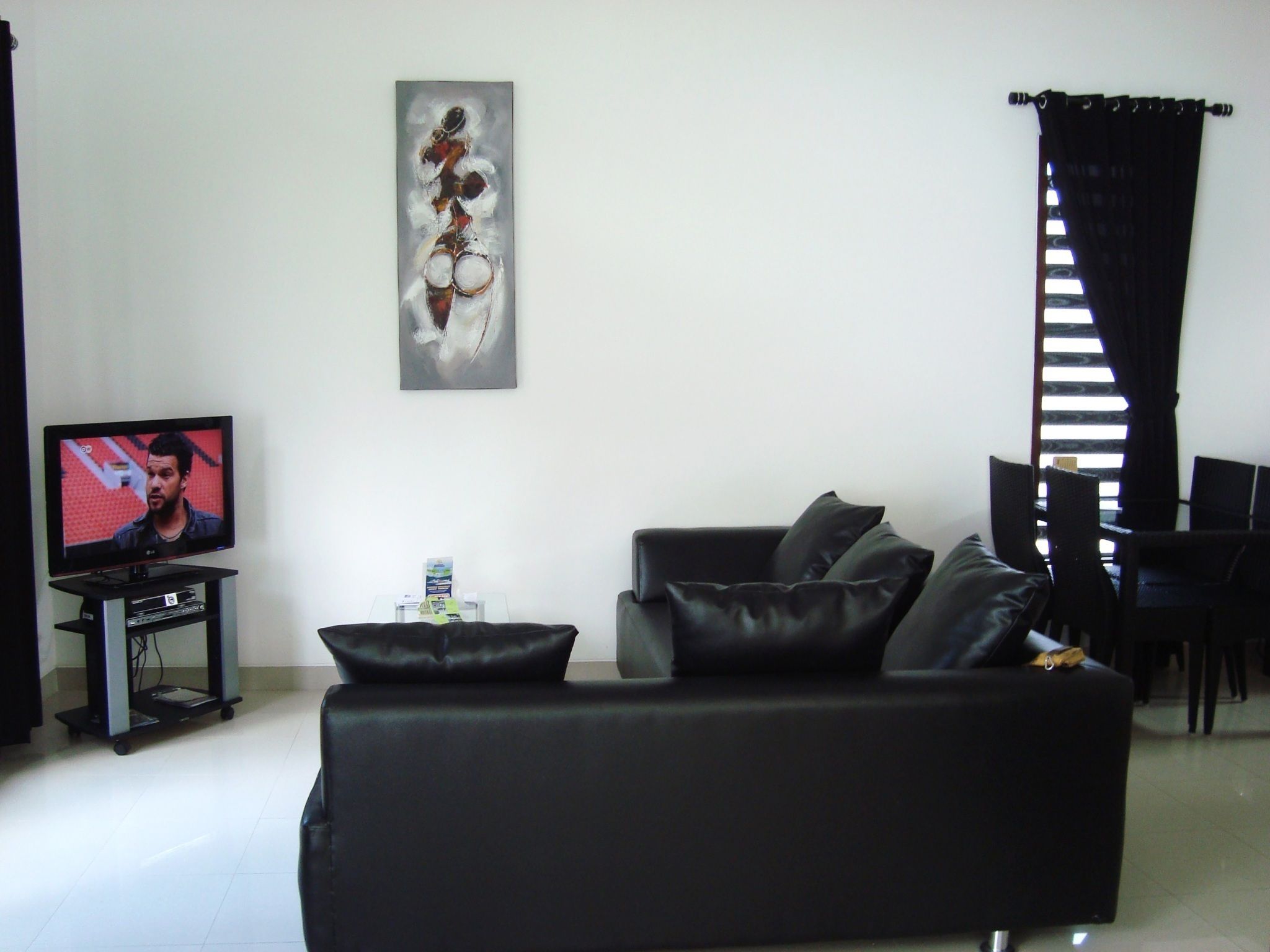 A living room with a black sofa, a flat-screen TV, a painting on the wall, and a window with black curtains.