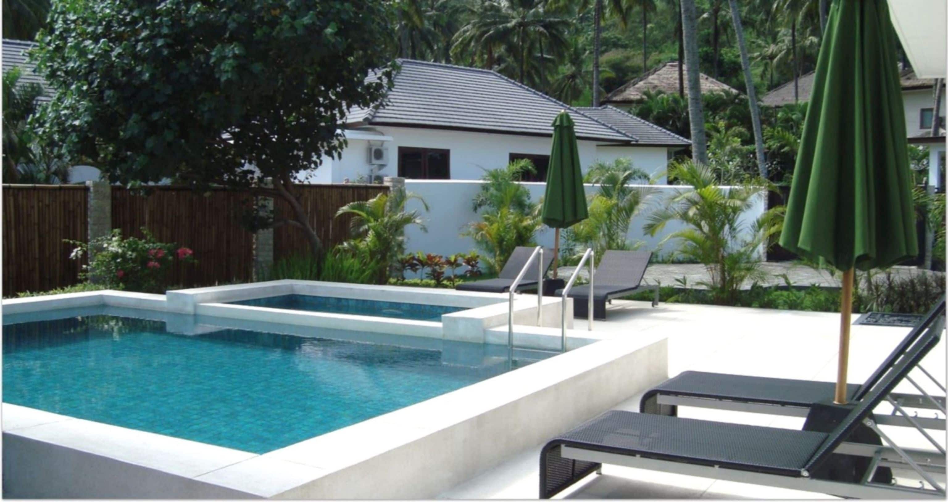 A rectangular swimming pool with two separate sections, surrounded by a white wall and a wooden fence. There are two lounge chairs and a green umbrella.