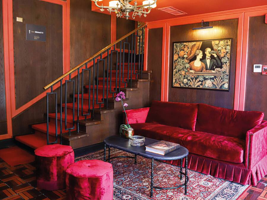 Una habitación con un sofá de terciopelo rojo, una mesita con un libro, una escalera y un cuadro en la pared.