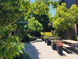 Un patio empedrado con plantas en macetas y bancos, rodeado de exuberante follaje y árboles.