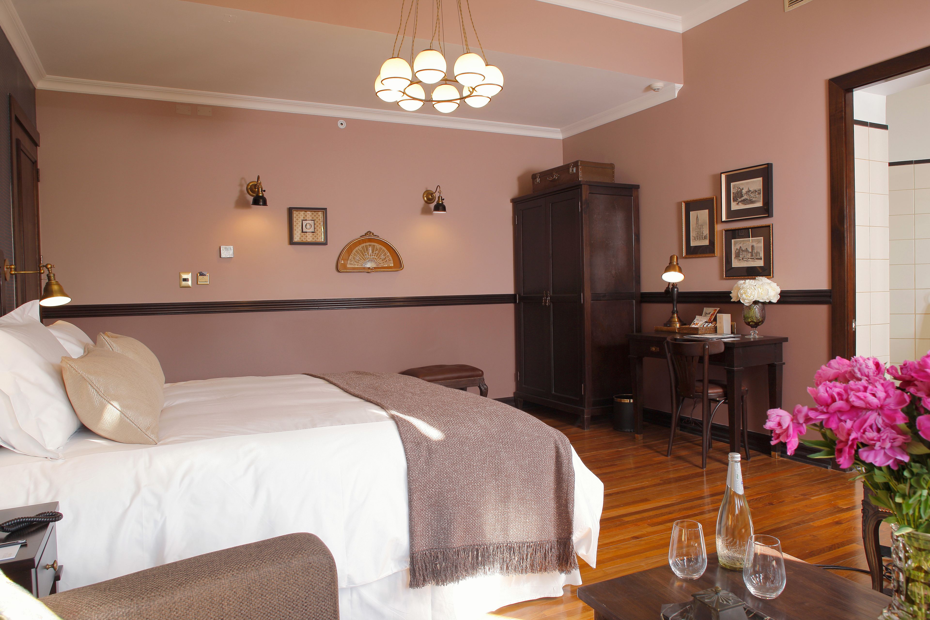 Una habitación de hotel con una cama, mesita de noche y un escritorio con una silla. Hay cuadros en la pared y un florero con flores en el escritorio.