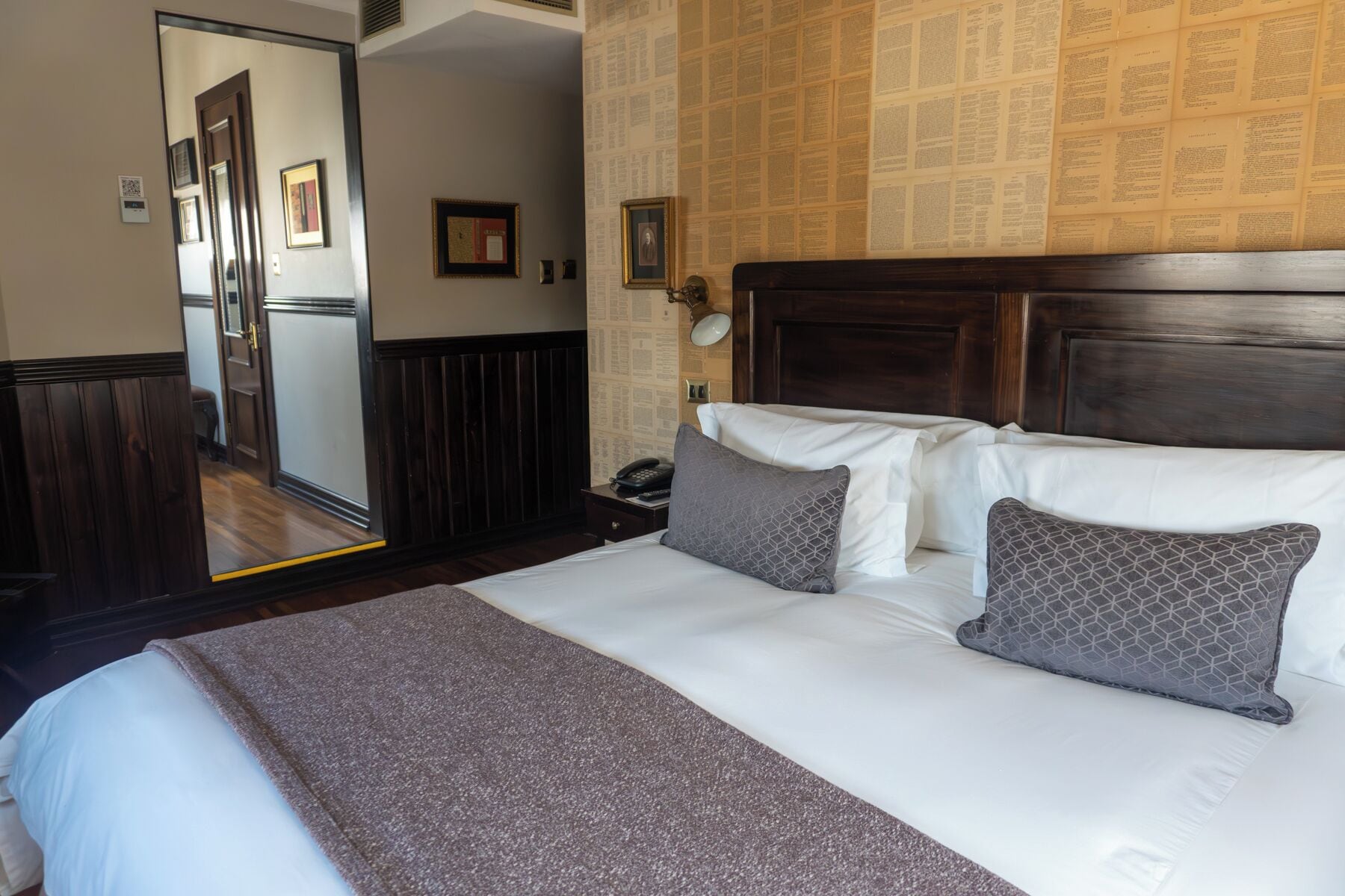 A hotel room with a large bed, a wooden headboard, a mirror, and a wall with newspaper clippings.