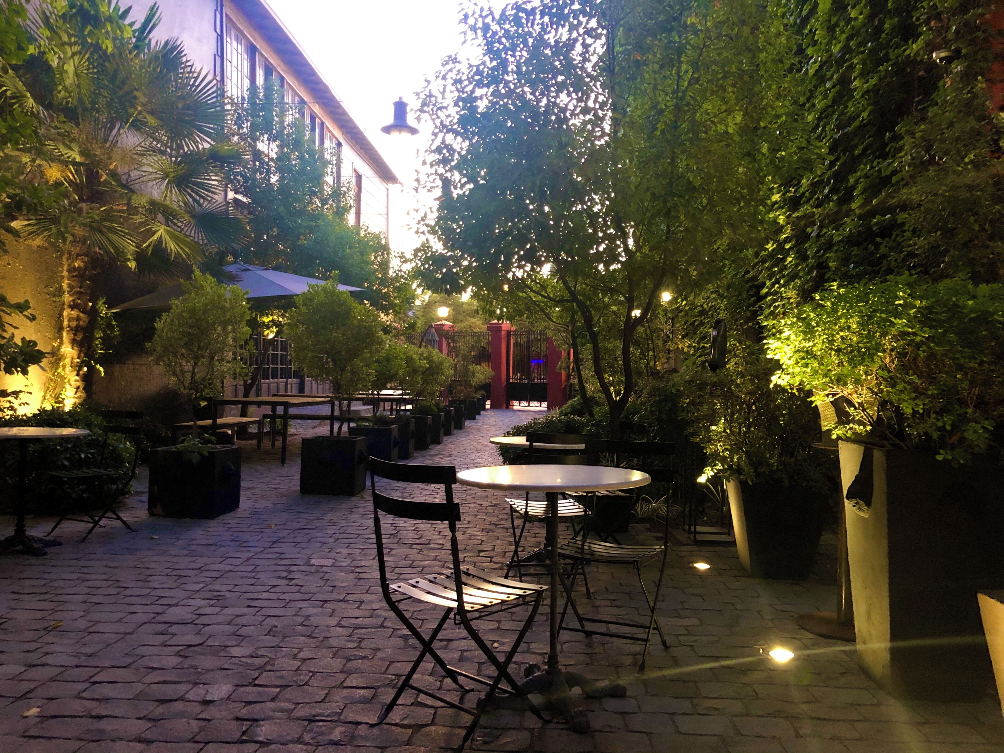 An outdoor patio area with tables and chairs, surrounded by greenery and a building in the background.