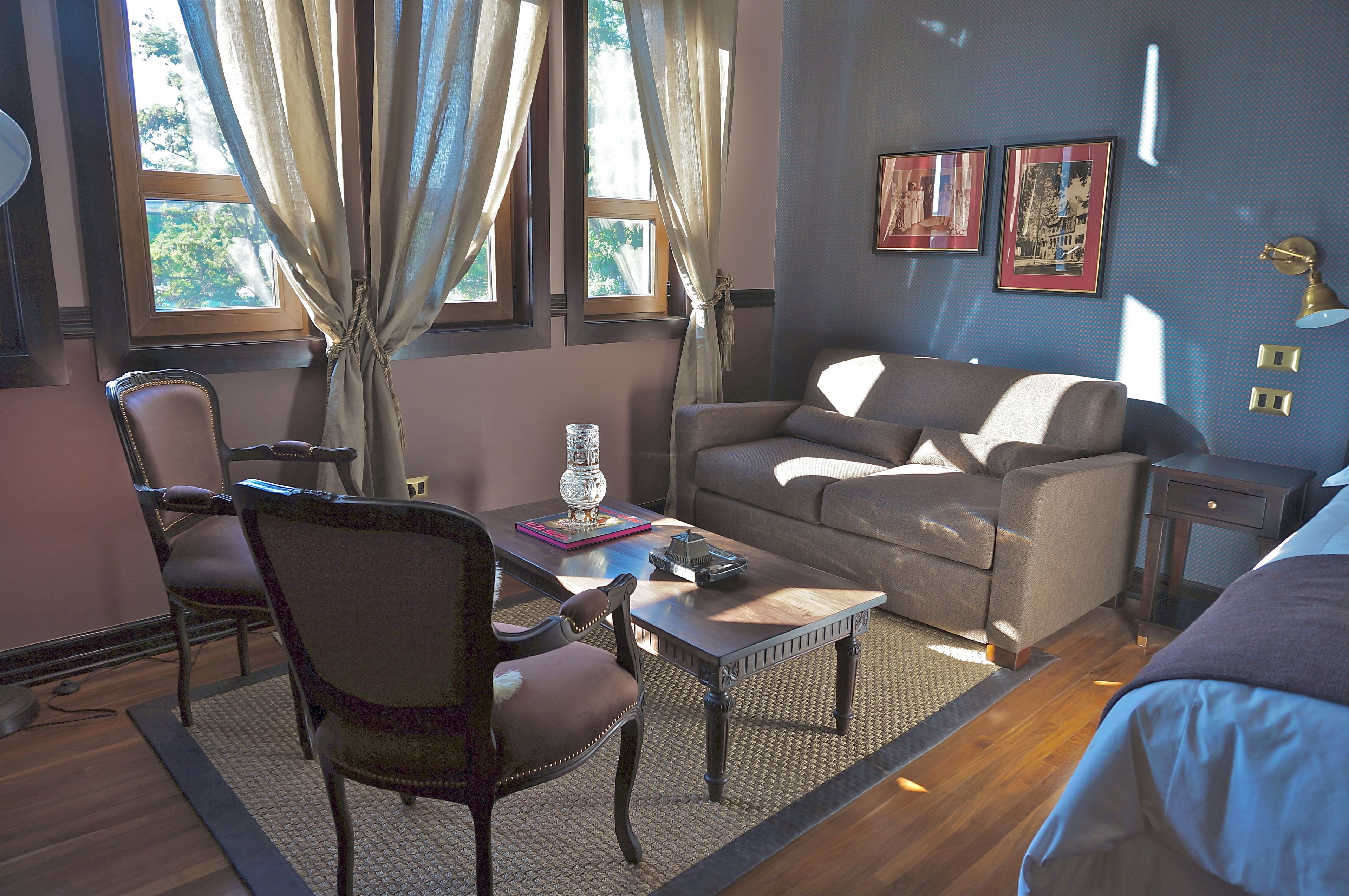 Una habitación acogedora con sofá, sillón, mesa de centro y lámpara. Hay dos ventanas con cortinas, una alfombra y un cuadro en la pared.