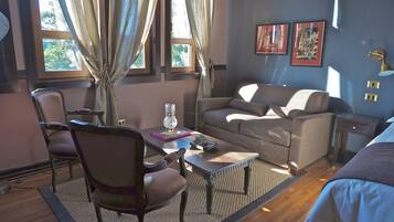 Una habitación acogedora con sofá, sillón, mesa de centro y lámpara. Hay dos ventanas con cortinas, una alfombra y un cuadro en la pared.
