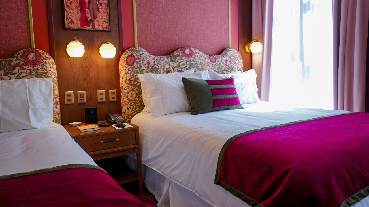 A hotel room with two beds, each with a floral headboard and white bedding. There are bedside tables with lamps and a clock.
