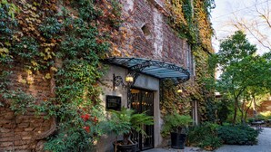 Un bâtiment à la façade en briques, aux murs recouverts de lierre et à la grille en fer forgé. Des plantes en pot et un chemin pavé de pierres mènent à l’entrée.