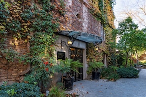 A building with a brick facade, ivy-covered walls, and a wrought iron gate. There are potted plants and a cobblestone path leading to the entrance.