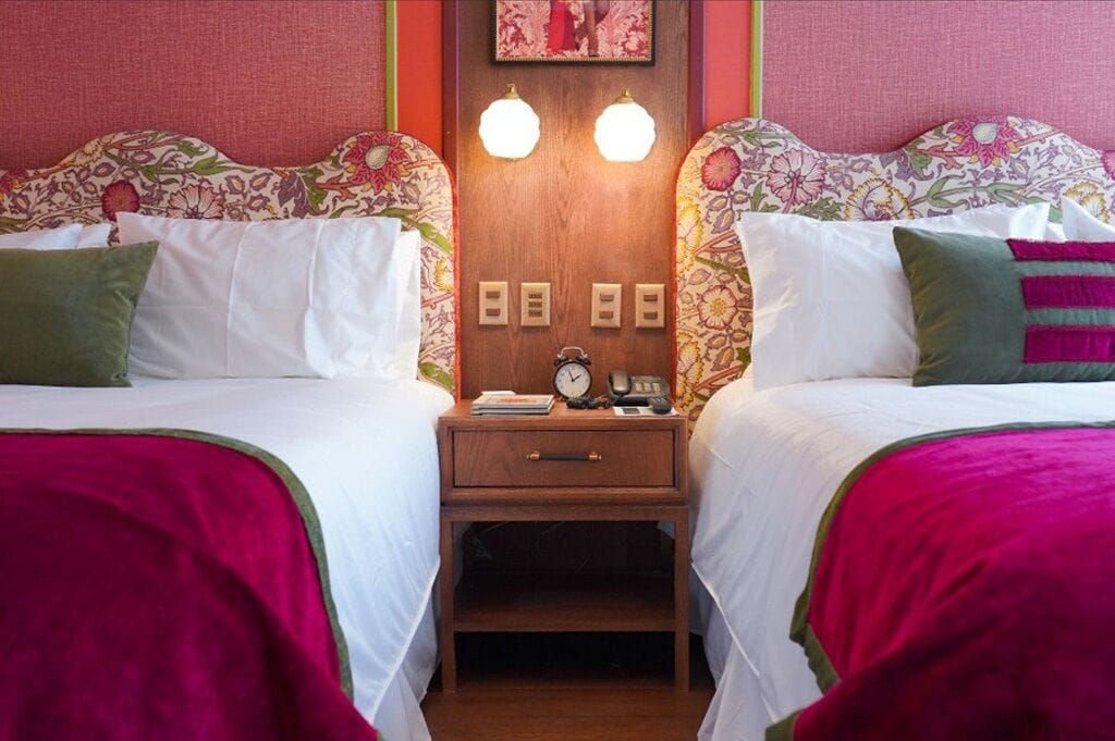 A hotel room with two beds, each with floral headboards, white bedding, and green pillows. There is a wooden nightstand with a clock and a small table between the beds.