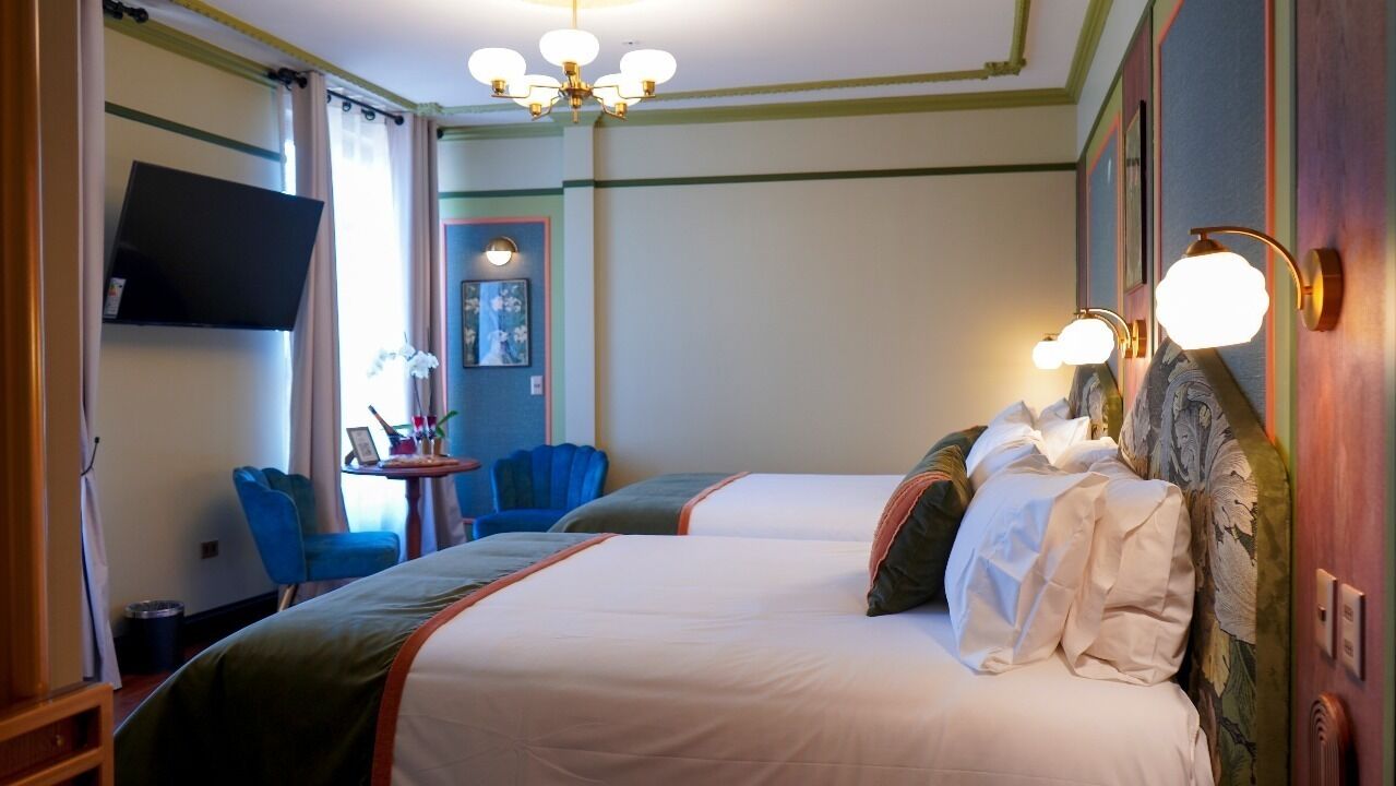 A hotel room with two beds, a TV mounted on the wall, a small table with chairs, and a chandelier.