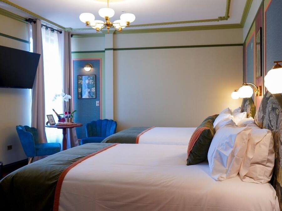 A hotel room with two beds, a TV mounted on the wall, a small table with chairs, and a chandelier.