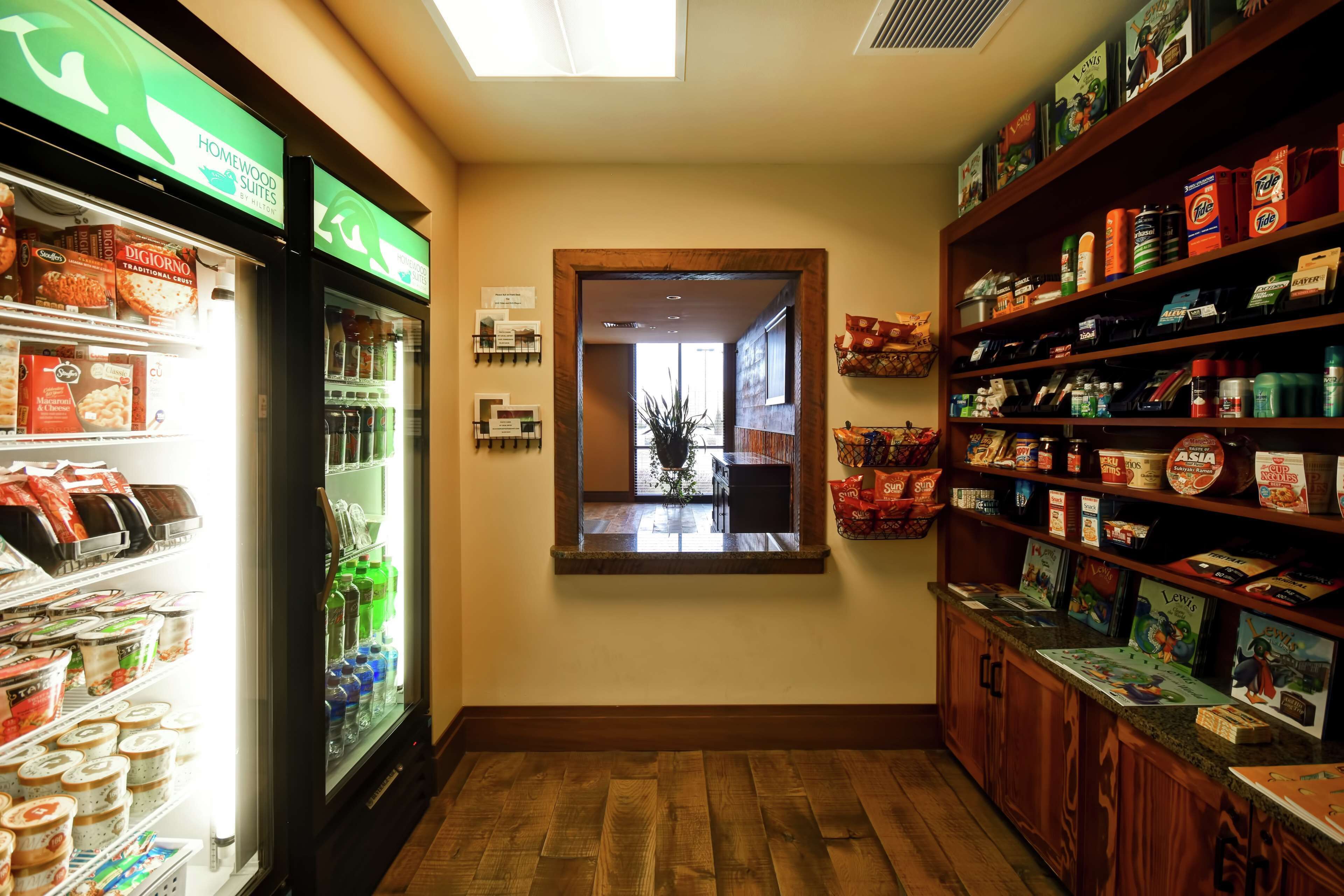Un pasillo con un refrigerador lleno de alimentos, un estante de madera con aperitivos y una ventana con vista al exterior.