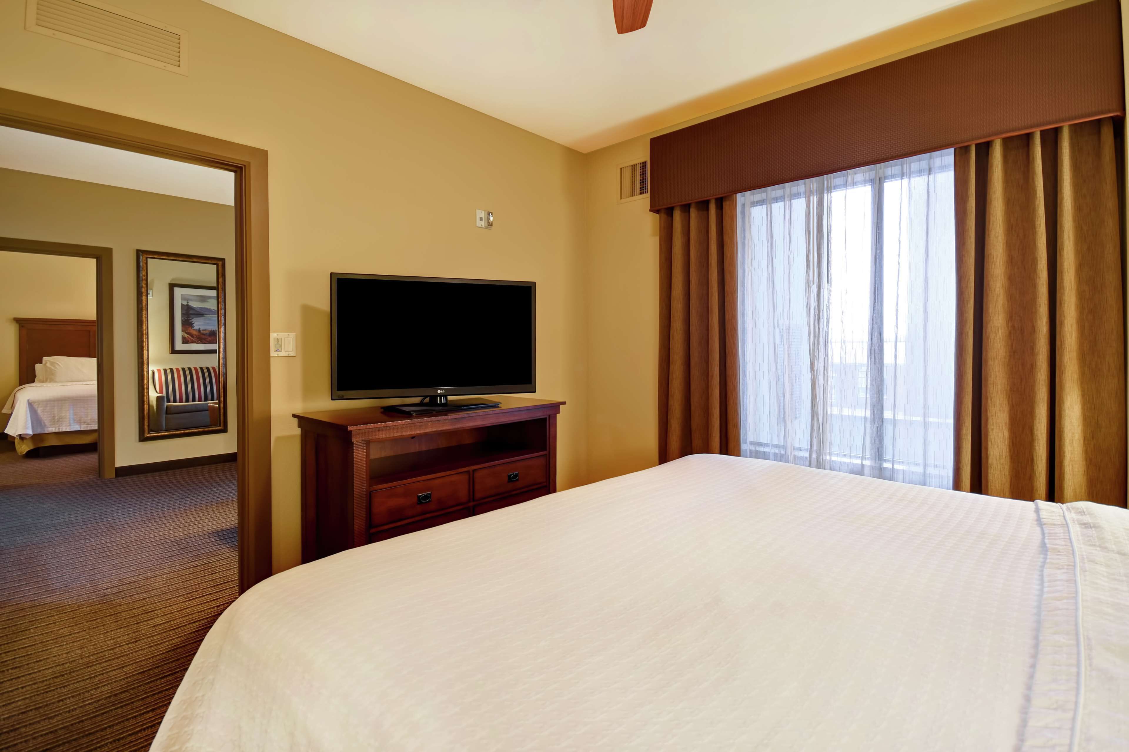 Habitación de hotel con una cama grande, un televisor de pantalla plana sobre un soporte de madera, una ventana con cortinas y una puerta que da a otra habitación.