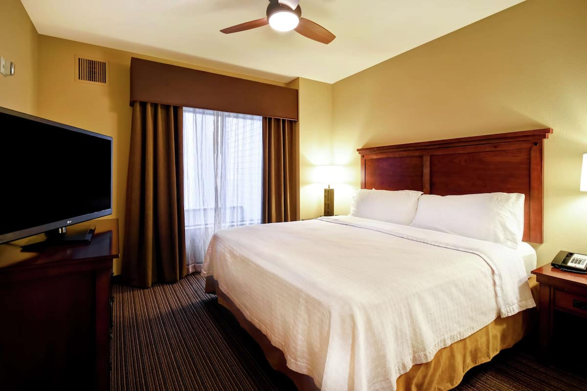 A hotel room with a large bed, a flat-screen TV, a ceiling fan, and a bedside table with a lamp.