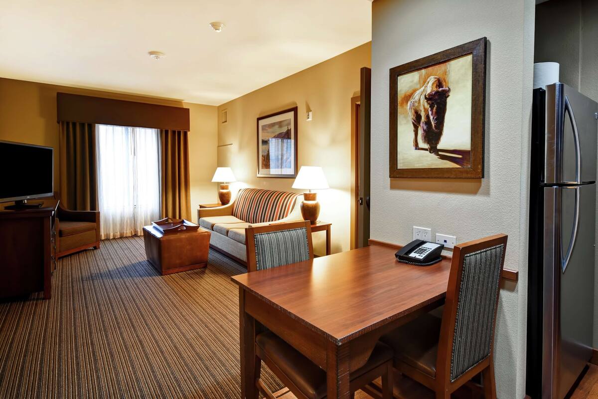 A hotel room with a flat-screen TV, a sofa, a dining table with chairs, a refrigerator, and a painting on the wall.