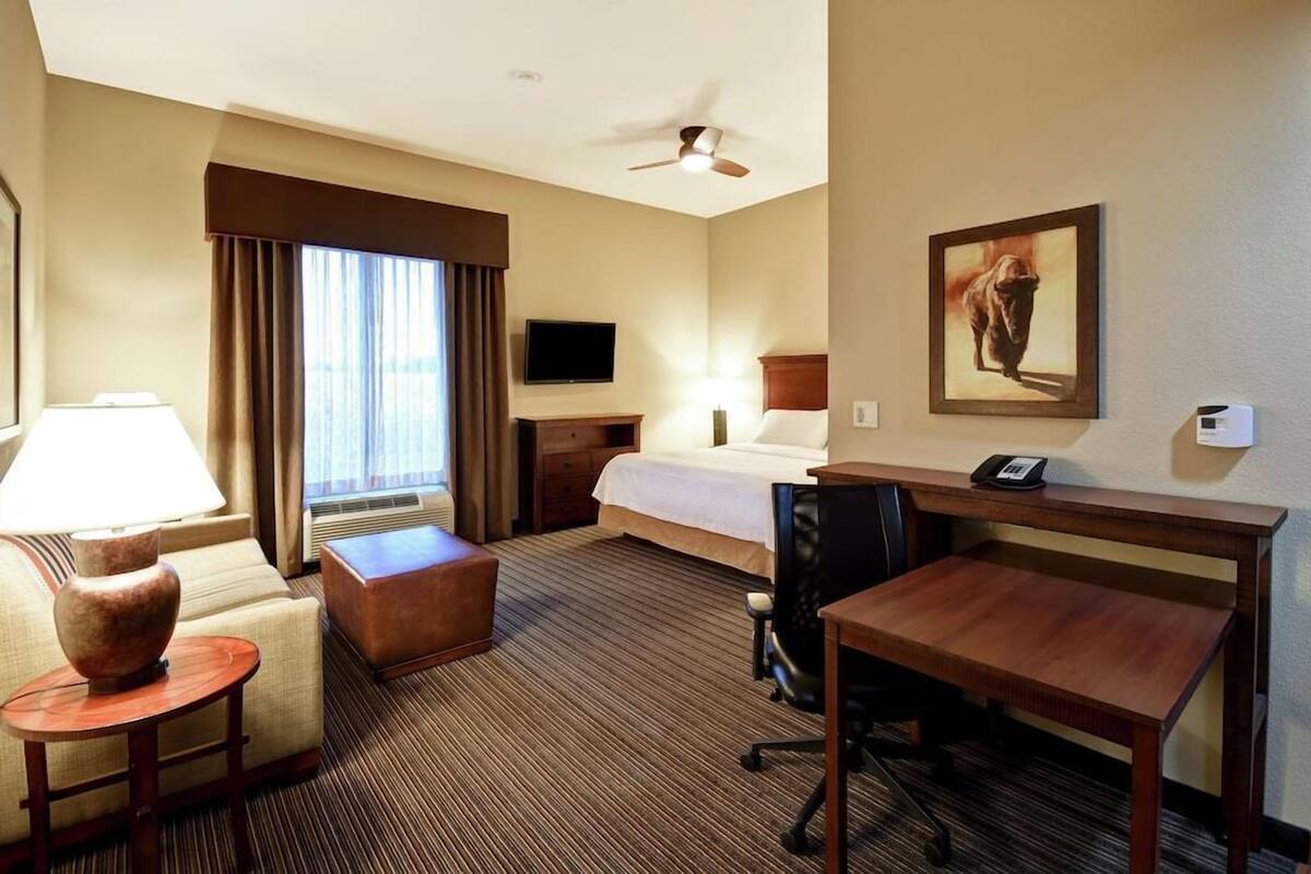 A hotel room with a bed, desk, chair, lamp, and a painting on the wall.