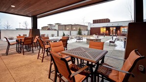 An outdoor patio with wooden tables and chairs, a stone fire pit, and a view of a building in the background.