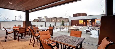 Un patio al aire libre con mesas y sillas de madera, un hogar de piedra y vistas a un edificio al fondo.
