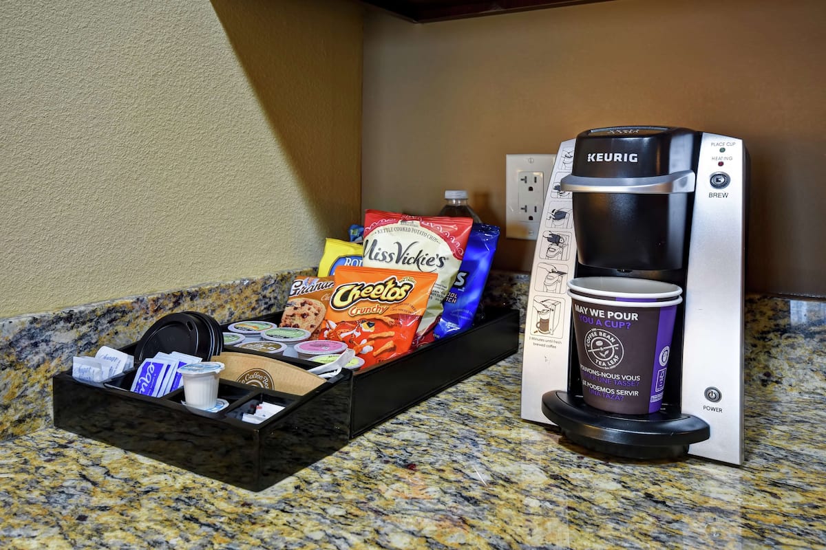 A coffee maker with a cup of coffee, a coffee pod holder, and a snack tray with chips and a drink.