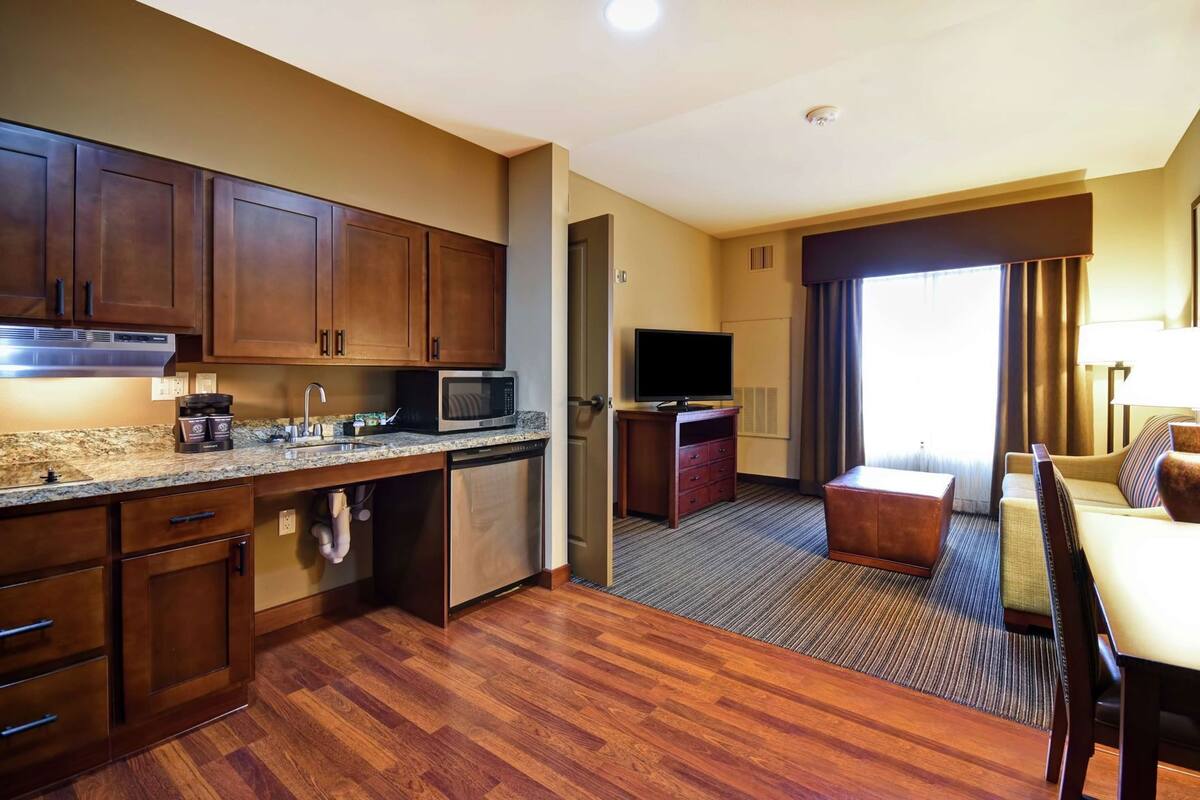 A hotel room with a kitchenette, a television, a sofa, and a window with curtains.