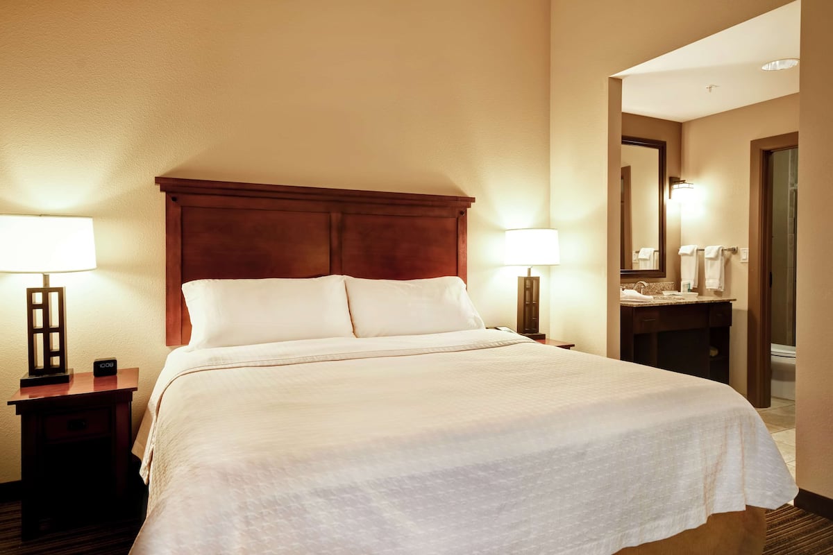 A hotel room with a large bed, two bedside lamps, a nightstand, a mirror, and a bathroom visible through an open door.