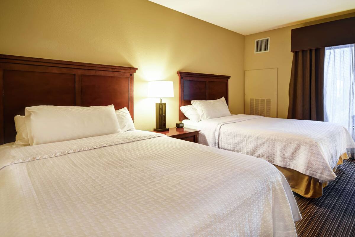 A hotel room with two beds, a nightstand with a lamp, and a window with curtains.