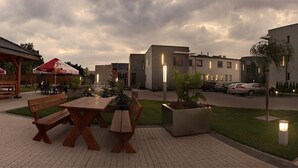 En udendørs patio med træbænke og borde, et overdækket siddeområde og moderne bygninger i baggrunden.