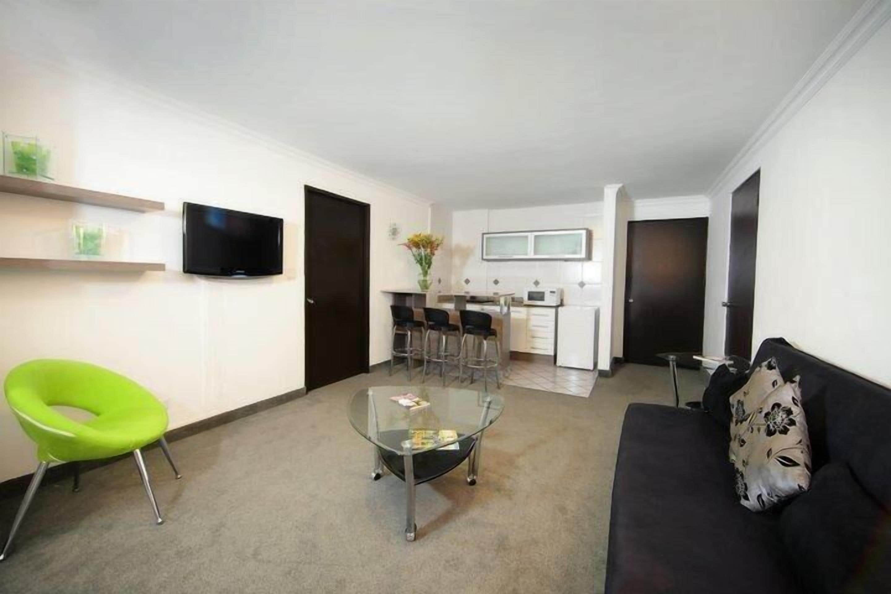 A compact living space with a glass coffee table, a green chair, a black sofa, and a kitchen area in the background.