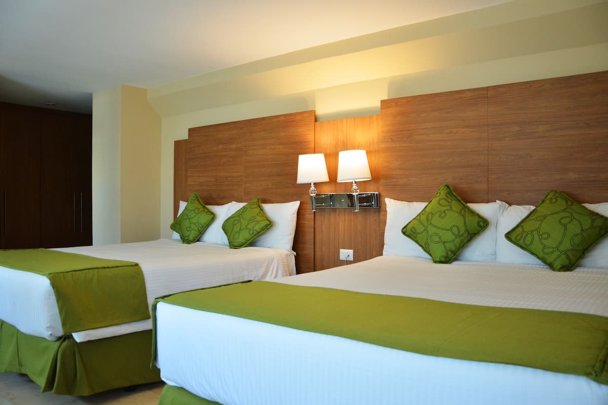A hotel room with two beds, each with green bedding and matching pillows. There are two wall-mounted lamps on the headboard.