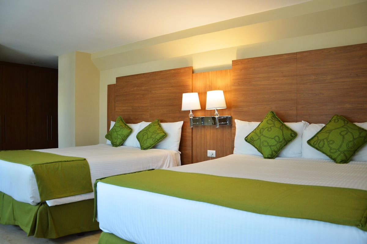 A hotel room with two beds, each with green bedding and matching pillows. There are two wall-mounted lamps above the beds.