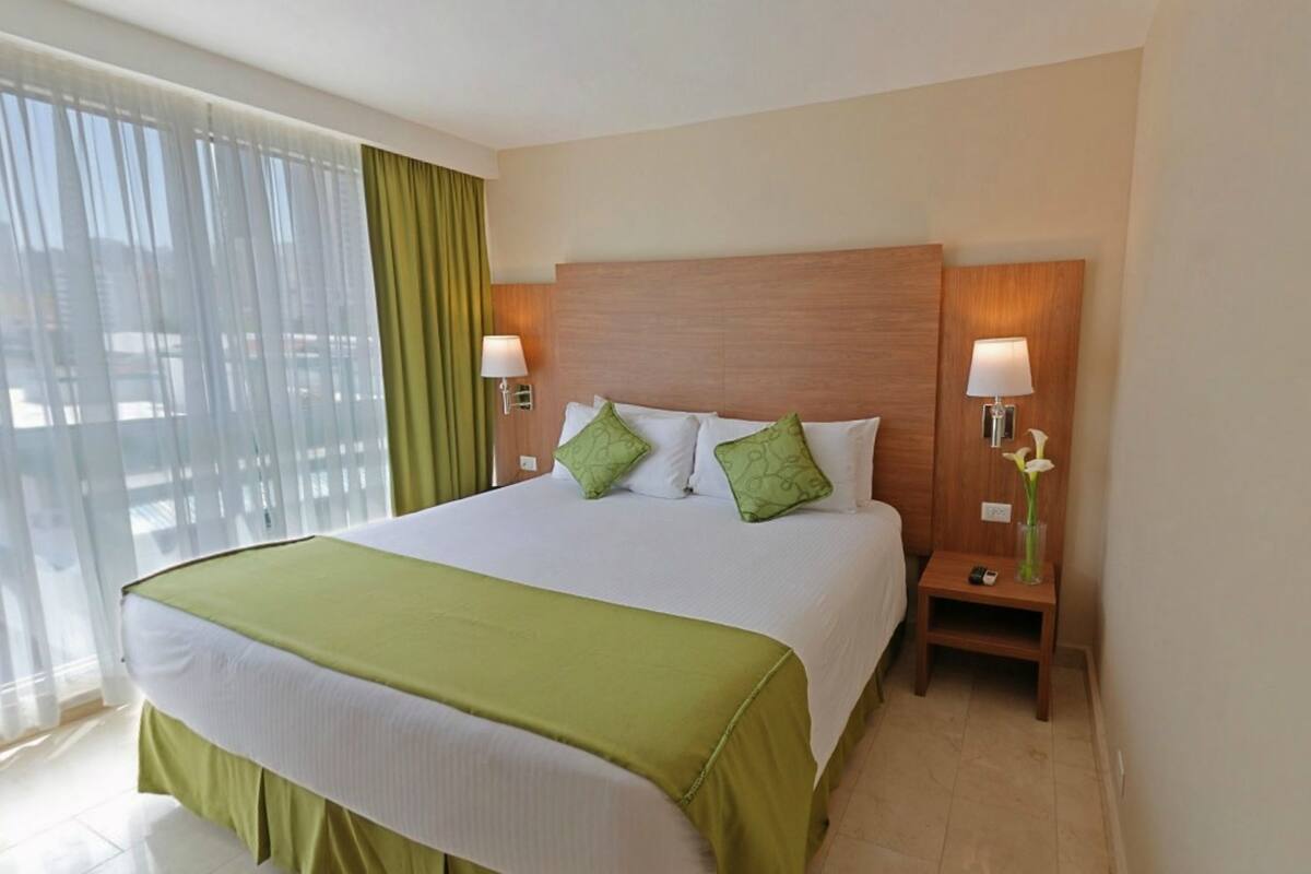 A hotel room with a large bed, green curtains, two bedside lamps, and a nightstand with a vase of flowers.